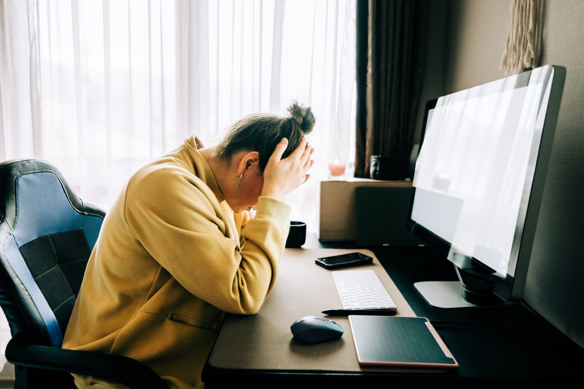 Woman working from home feeling stressed and frustrated, holding her head in her hands while sitting at her desk - stock photo