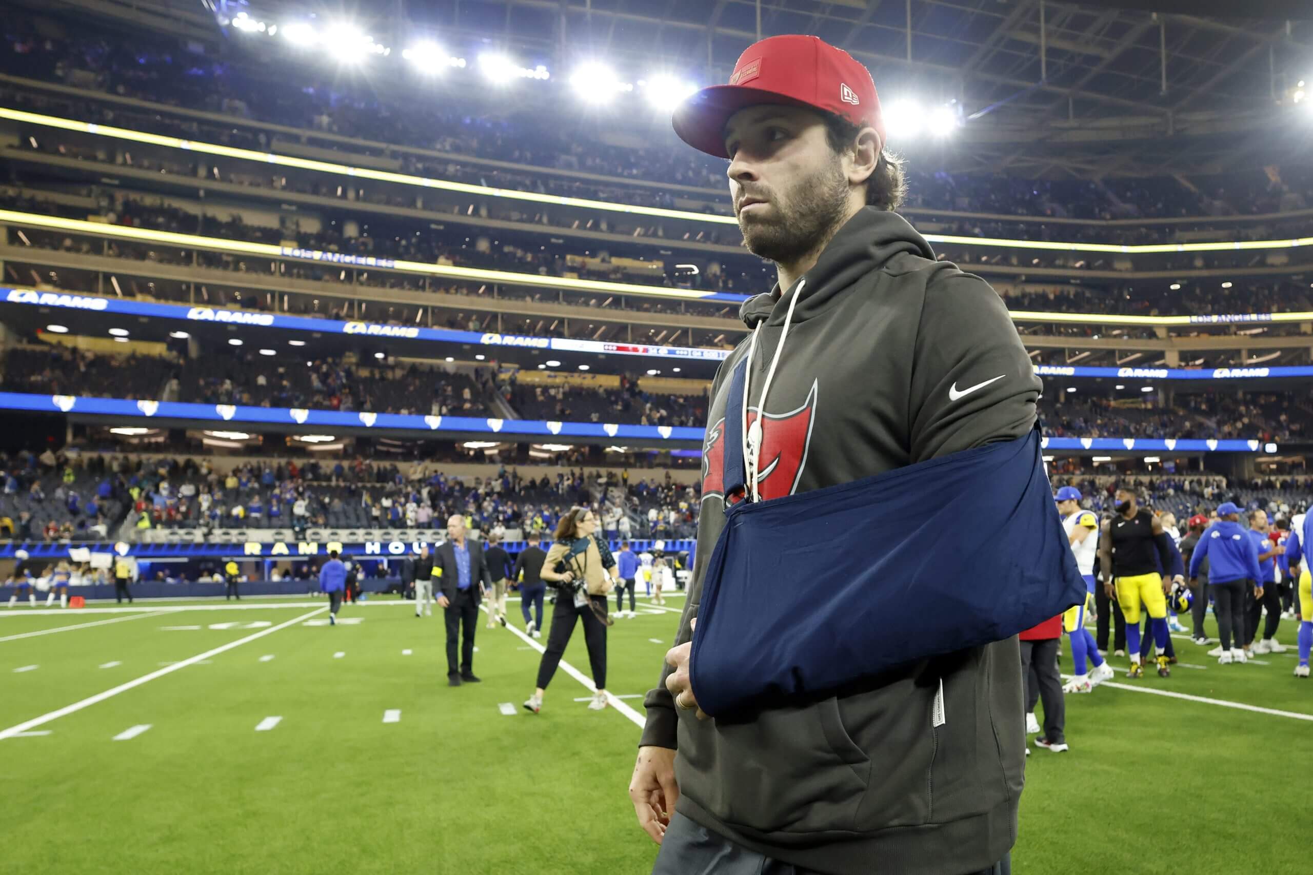 Baker Mayfield, wearing a gray hoodie and red hat, walks off the Rams' field with his left arm in a sling.