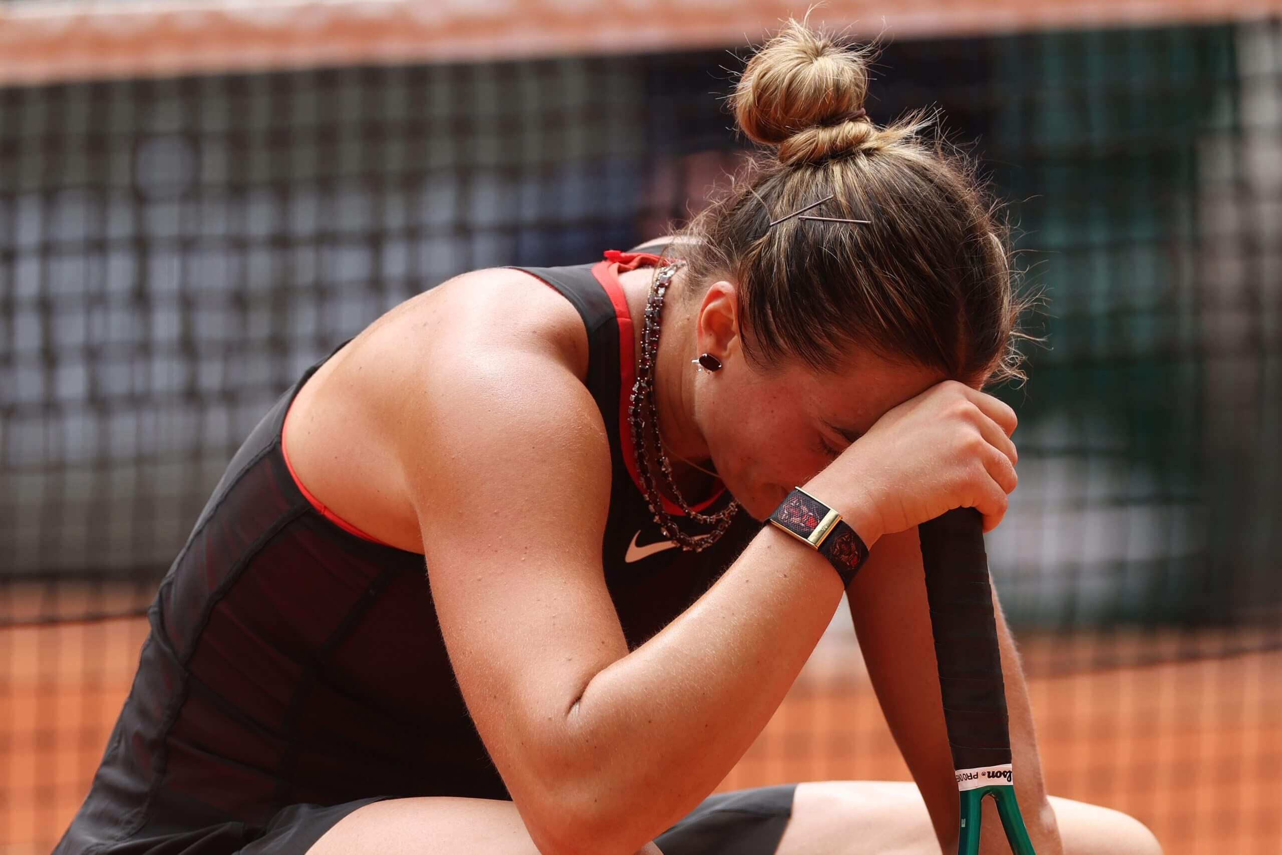 Aryna Sabalenka sits on her haunches, hand and head on top of the handle of her tennis racket.