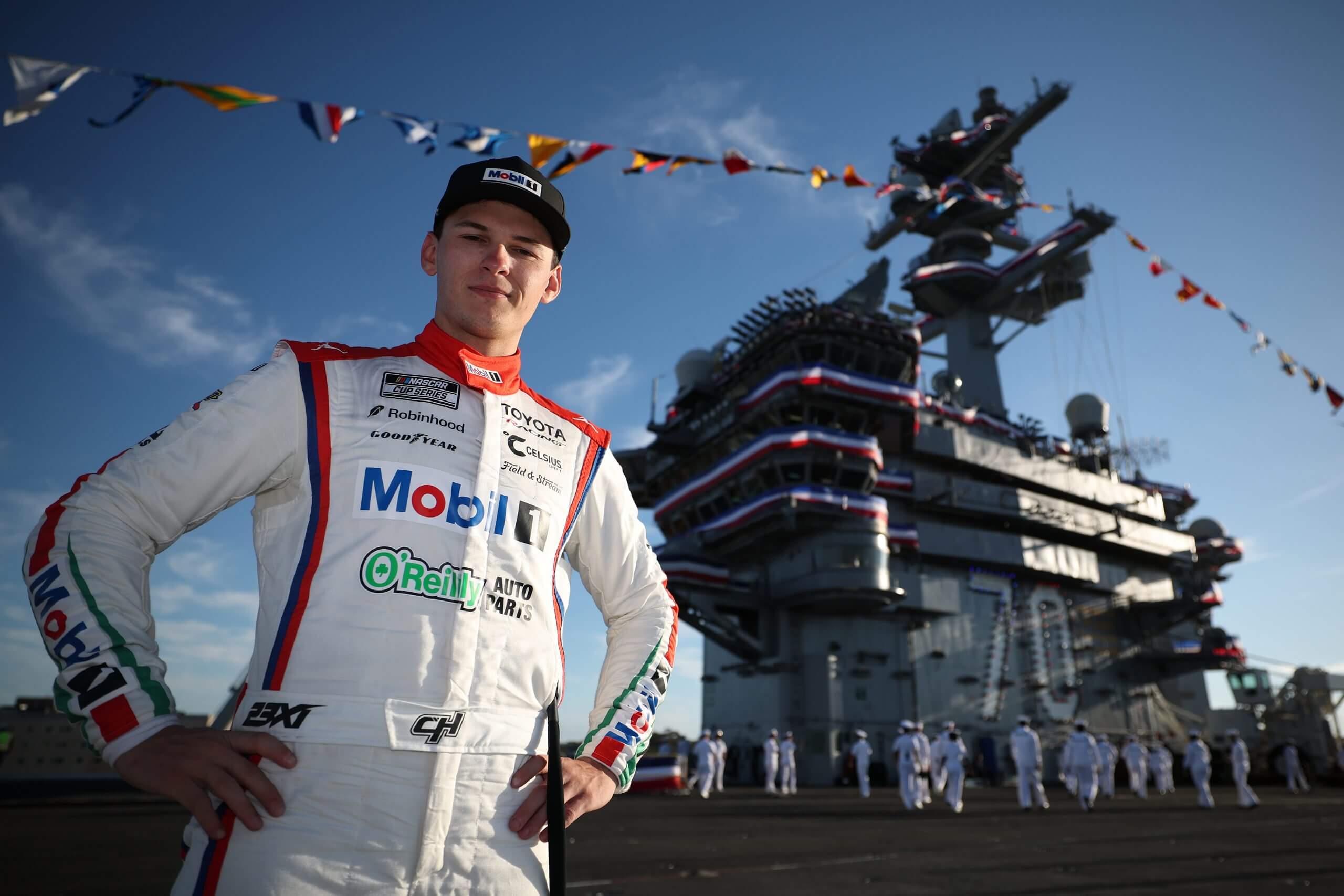 Corey Heim stands with his hands on his hips in front of a Navy ship with flag banners.