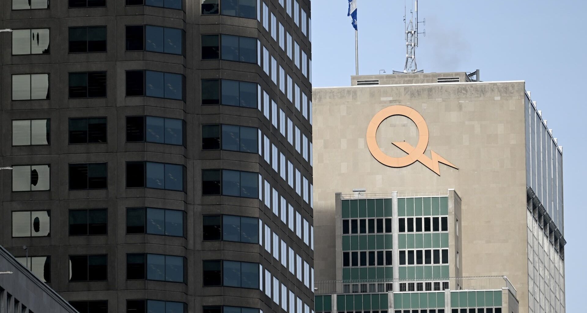 A cityscape featuring two tall buildings; the right one has a large orange "Q" logo and a Quebec flag atop. The sky is clear and blue.