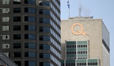 A cityscape featuring two tall buildings; the right one has a large orange "Q" logo and a Quebec flag atop. The sky is clear and blue.