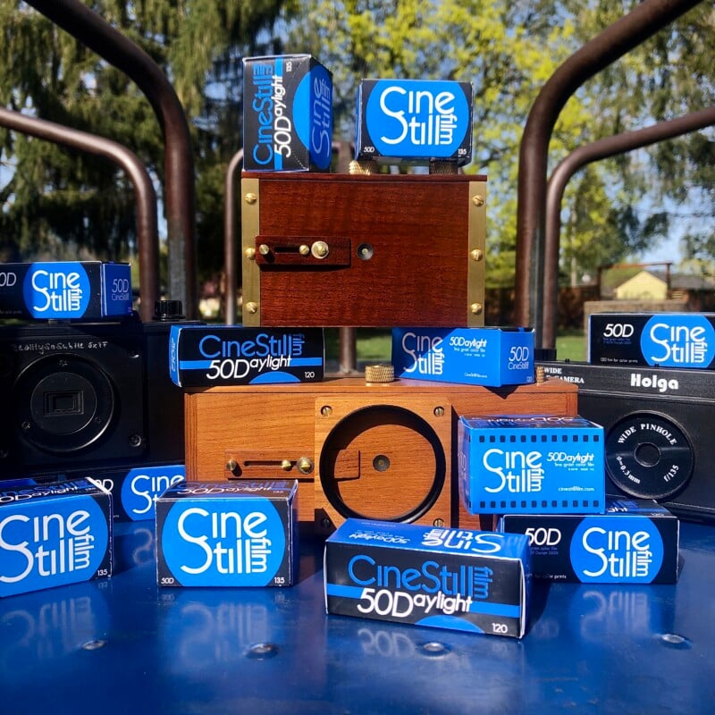 Several CineStill 50D Daylight film boxes are arranged among vintage and toy cameras outdoors, with trees and sunlight in the background.