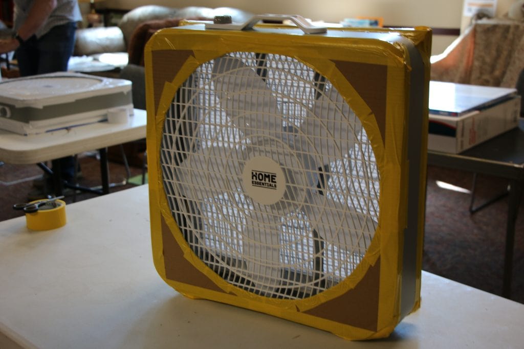 A DIY air cleaner made out of a box fan, a furnace filter and duct tape sits on a folding table. The face of the fan has a "shroud" made of cardboard. It blocks off the corners and edges of the fan to prevent air from flowing back into the fan