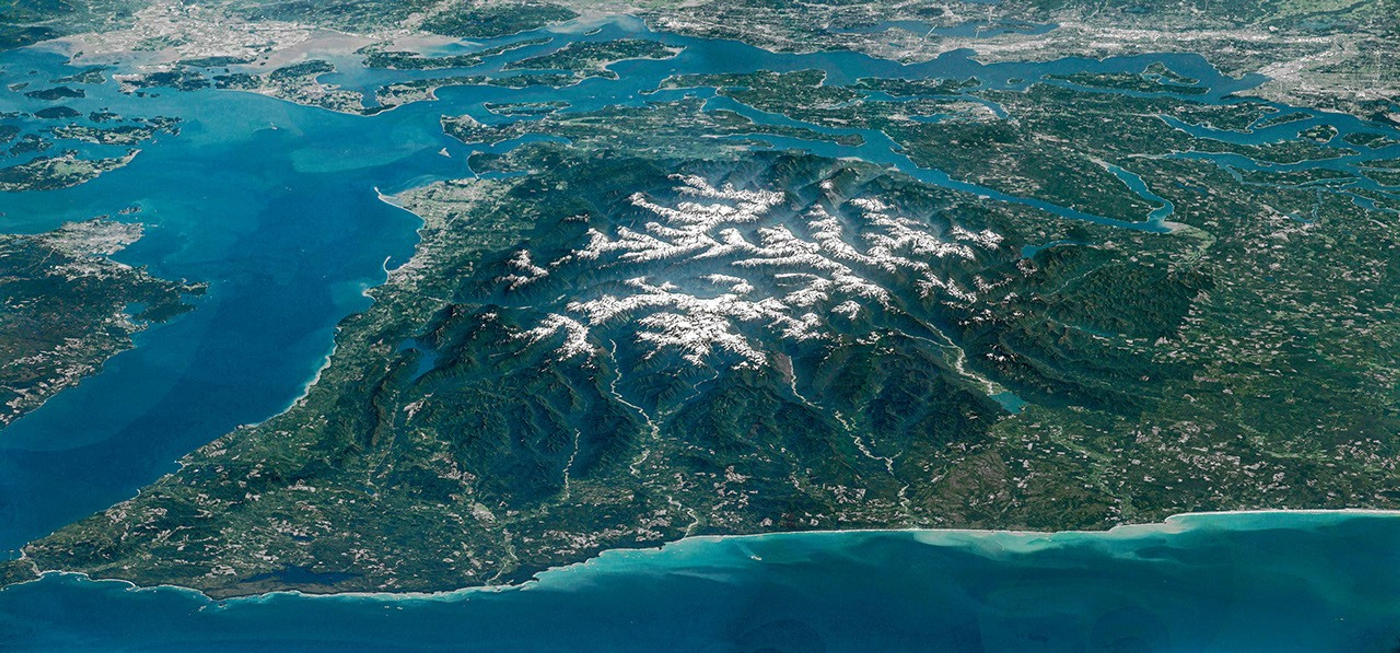The Olympic Peninsula, viewed at an angle from above, features snow-capped mountains surrounded by deep, forested river valleys. Islands in Puget Sound and developed areas including Seattle and Tacoma appear across the top of the photo.