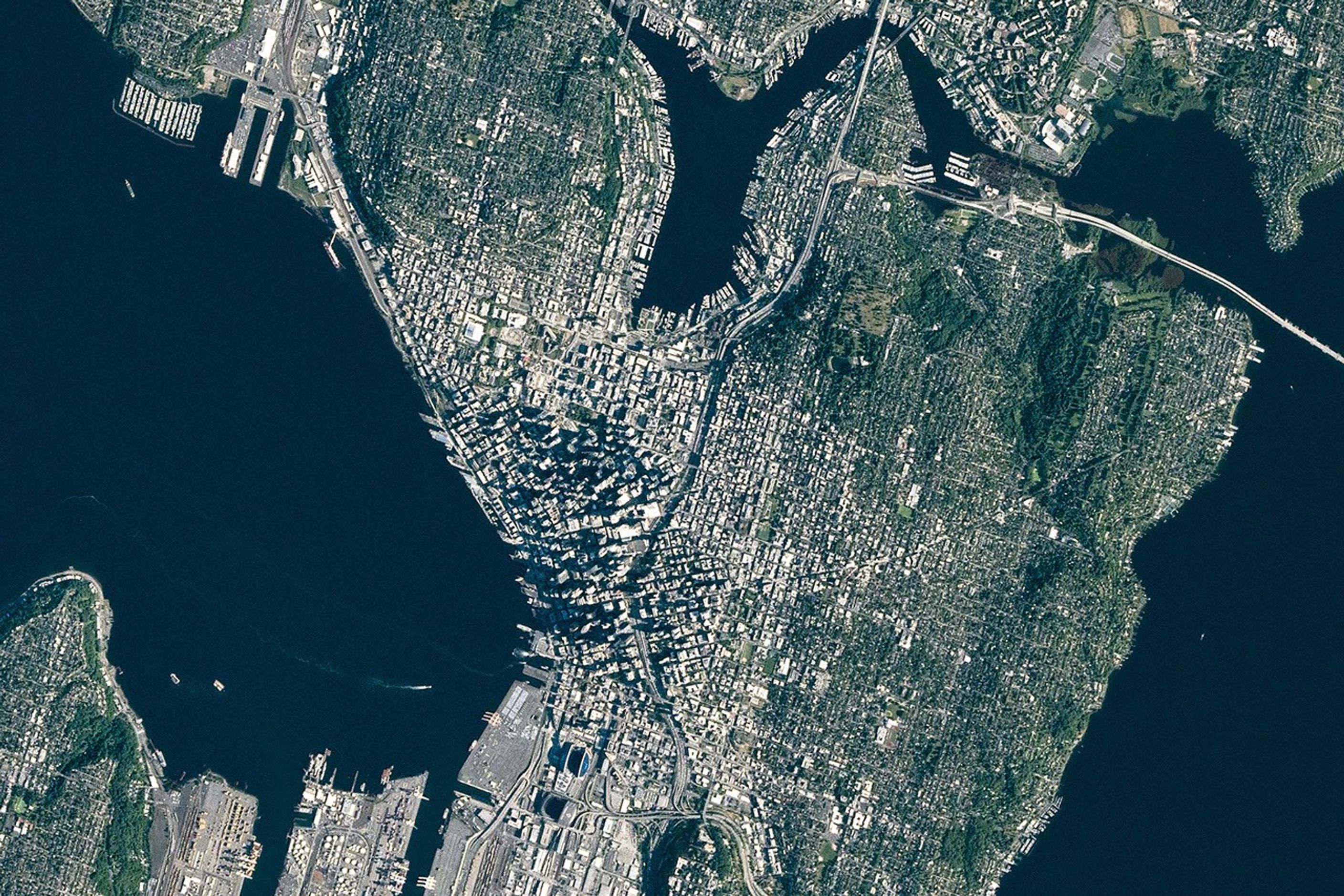 A top-down photo focuses on downtown Seattle, where tall buildings cast long shadows. Green neighborhoods and parks surround the dense urban development, and large waterbodies lie to the east and west.