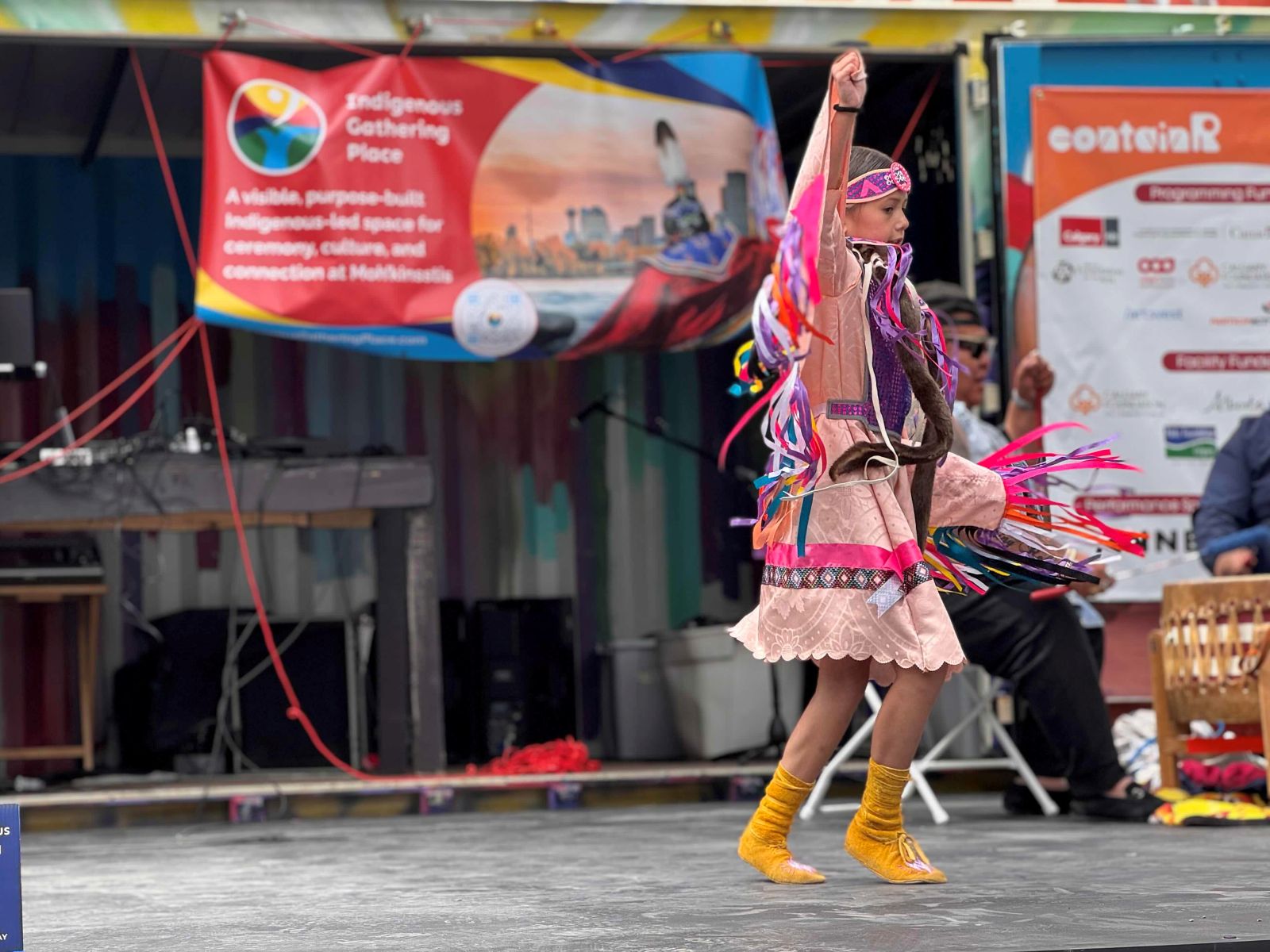 A girl in colourful pink Indigenous regalia dances onstage wearing yellow moccasins. Two men are seated onstage to the back right of the frame, drumming.