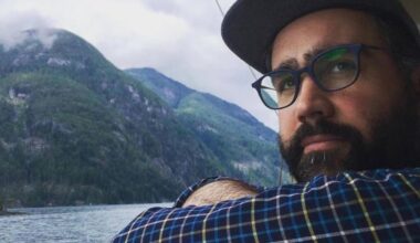 A light skin toned and bearded man wearing a baseball hat and a striped shirt looks on as a background of sea and hillside rolls by behind him.