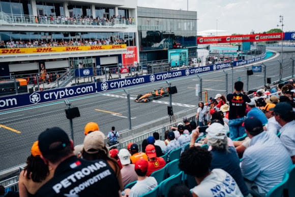 Crowded grandstand watches a race car speeding along a pit-lane street circuit with blue crypto.com banners.
