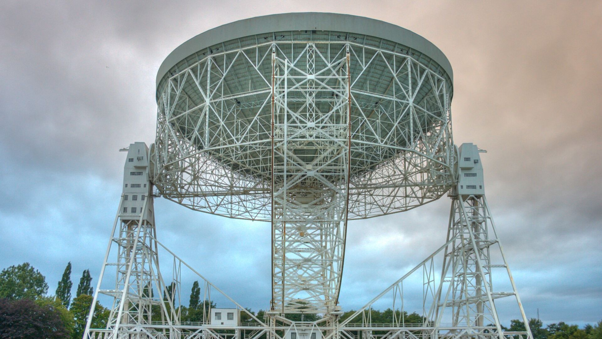 A photo of a white large structure with lots of poles holding it up. The poles conglomerate into some large stands that look like "legs" of the large cylinder structure that's raised above ground. This is outdoors.