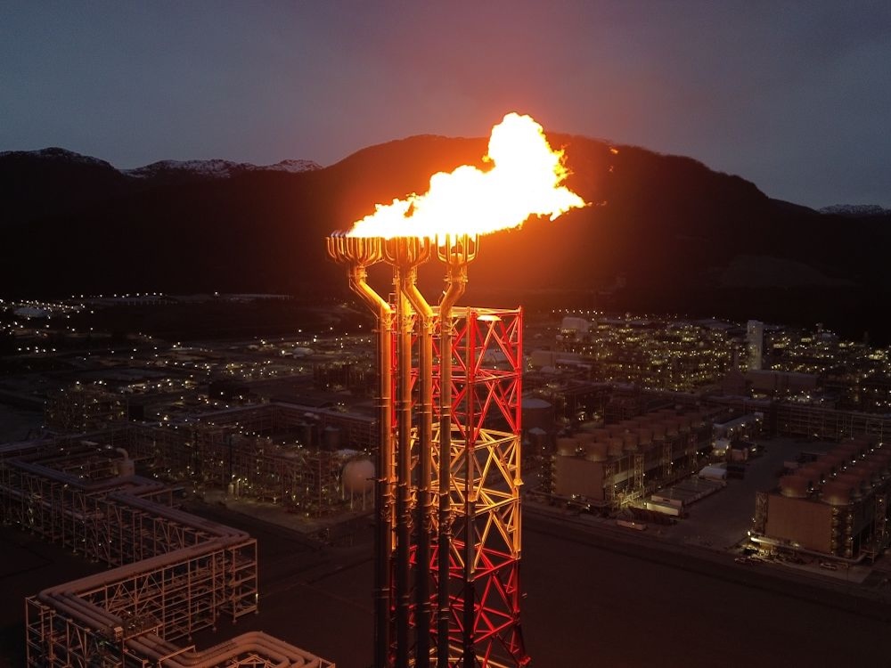 A large flame comes from the top of a metal tower in a mountainous area.