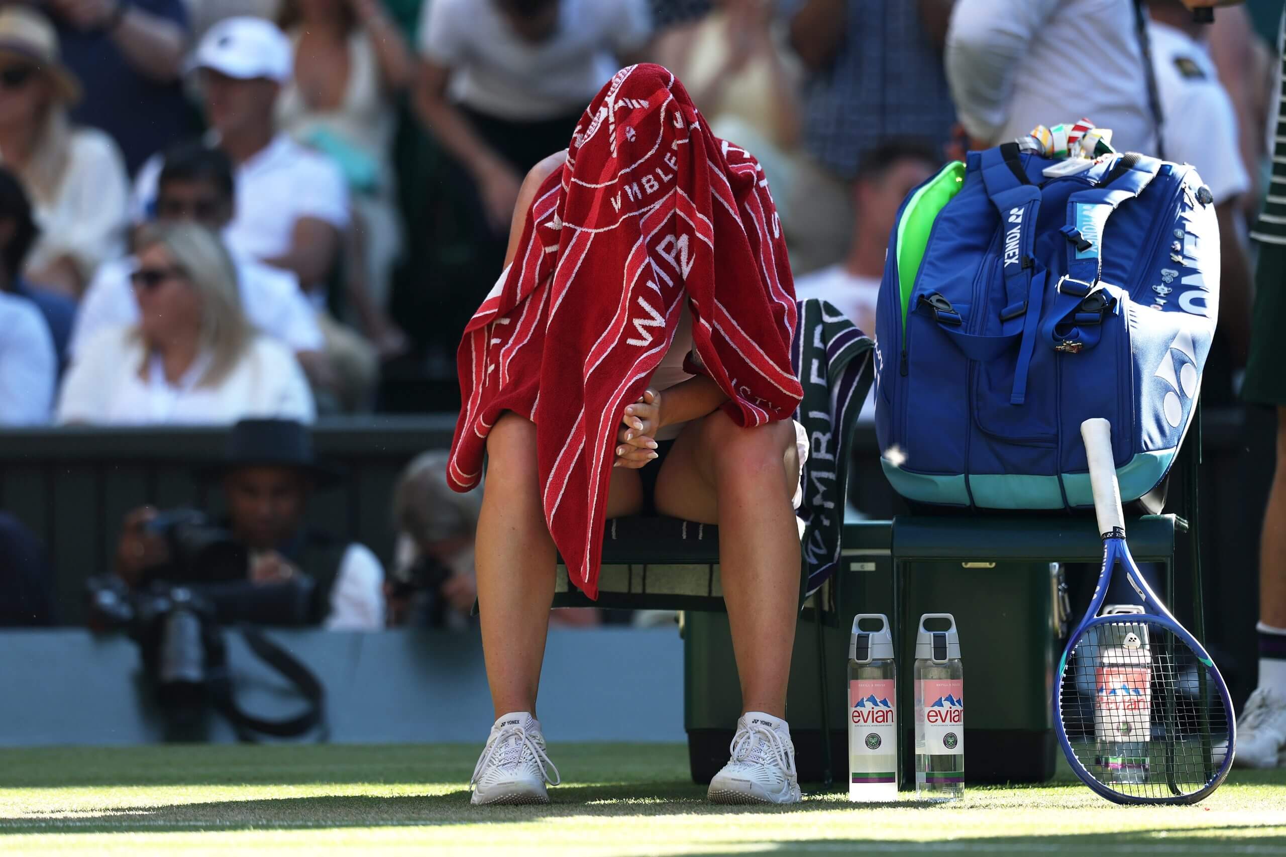 Linda Nosková sits with her head in a red towel at the side of a grass tennis court.