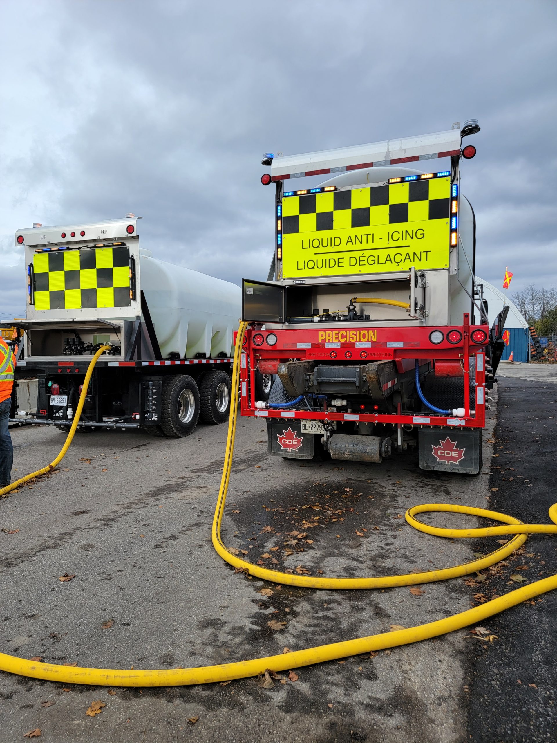 Two trucks with a yellow checkered sign on the backs. One truck says "Liquid Anti-Icing."