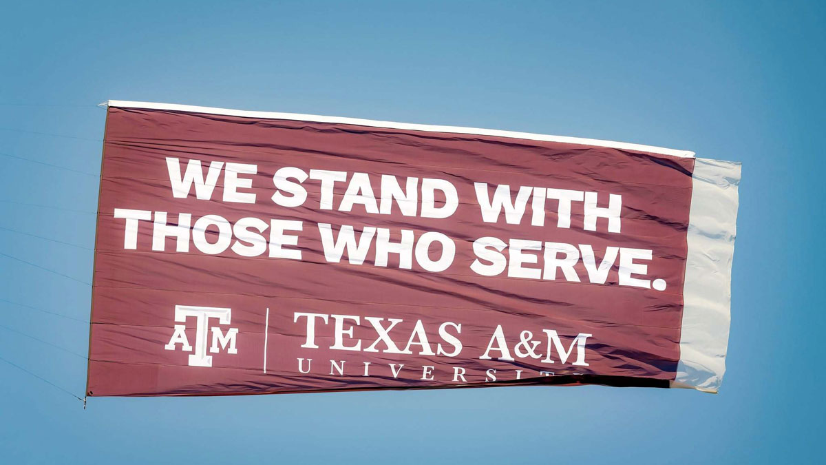 Banner flying over the Anduril 250 NASCAR race that says We Stand With Those Who Serve, followed by the Texas A&M logo