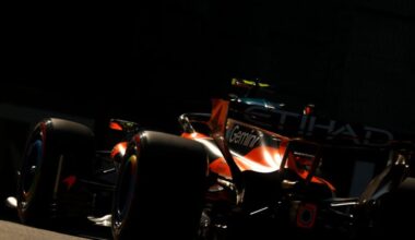 Rear three-quarter view of a red Formula 1 car on a race track at dusk, with large slick tires and rear wing visible behind the wheel arches and shadowy surroundings.
