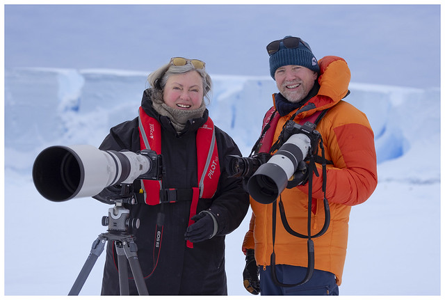 Photographers Sue Flood and Ian Dawson stand smiling together with long telephoto lenses in front of an Antarctic ice wall.