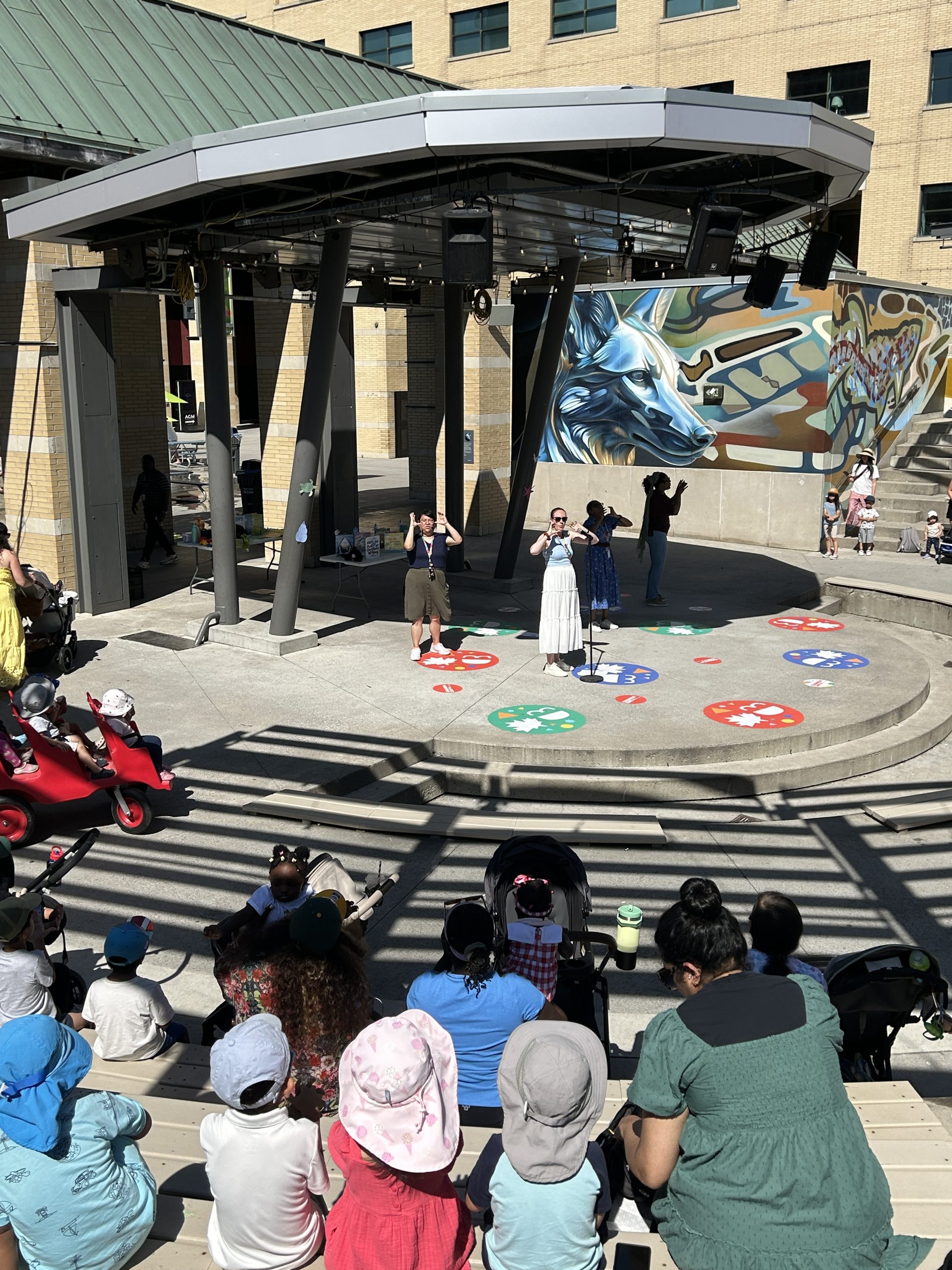 Community members attend an outdoor library storytime and performance at an amphitheatre. Presenters lead activities on stage while children and caregivers watch from the audience.