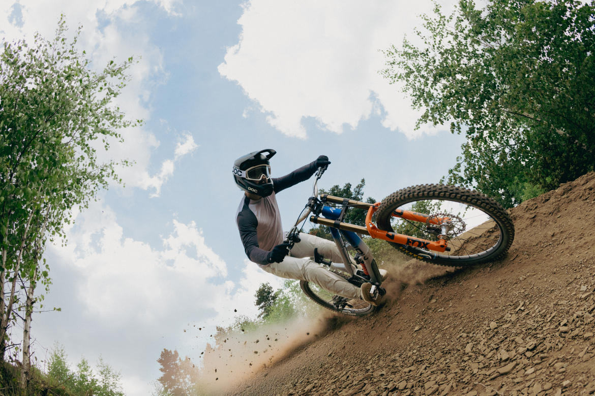 A mountain biker in protective gear leans into a turn on a dirt trail, kicking up dust as they navigate through a forested area under a partly cloudy sky.