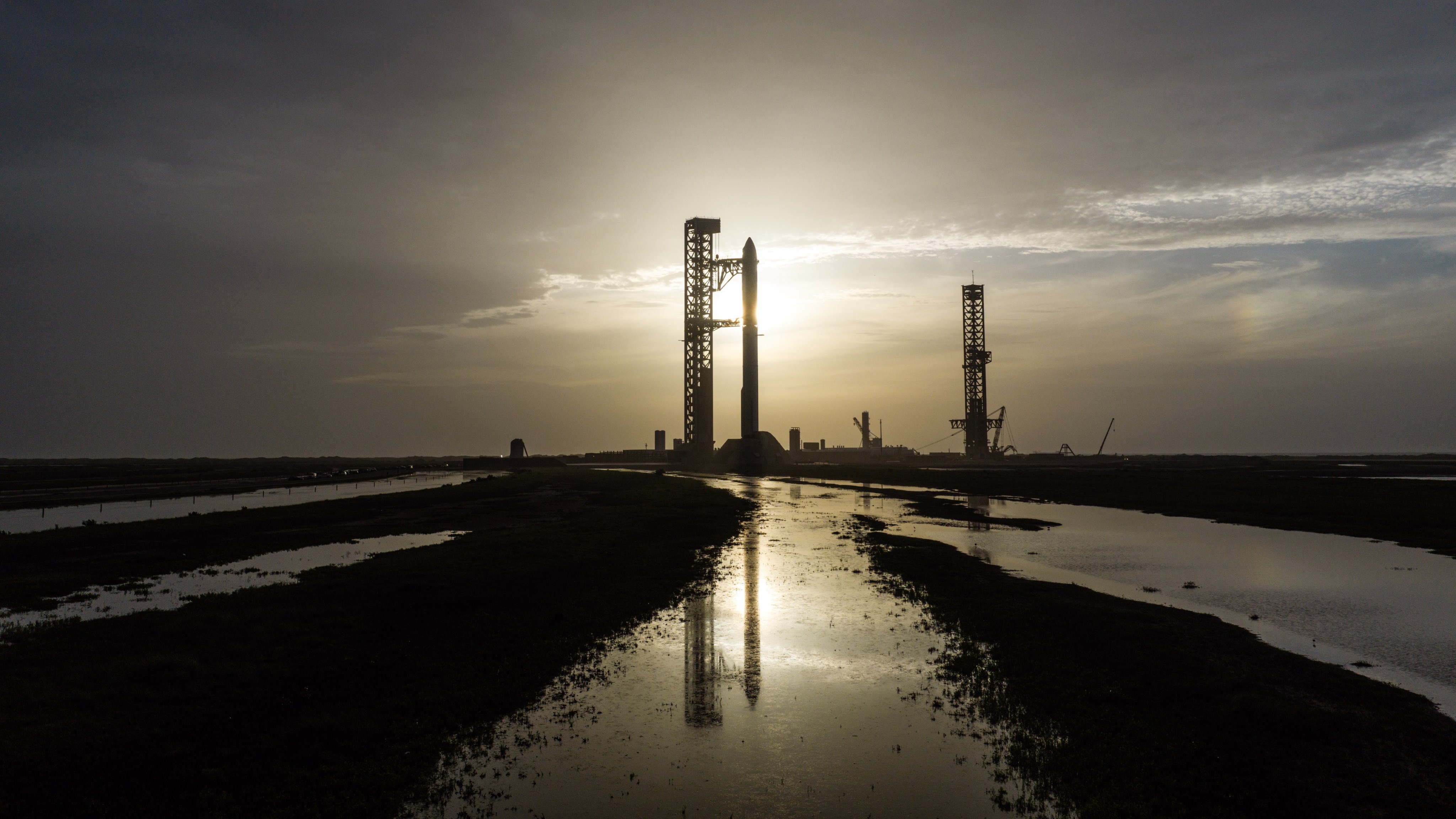 A giant steel rocket stands next to a launch tower silhouetted in front of the sun and reflected in thin pools of water in the foreground.