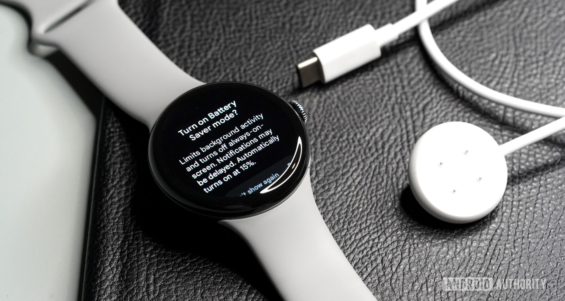 A Pixel Watch 3 display the Battery Saver mode screen rests alongside its charger.