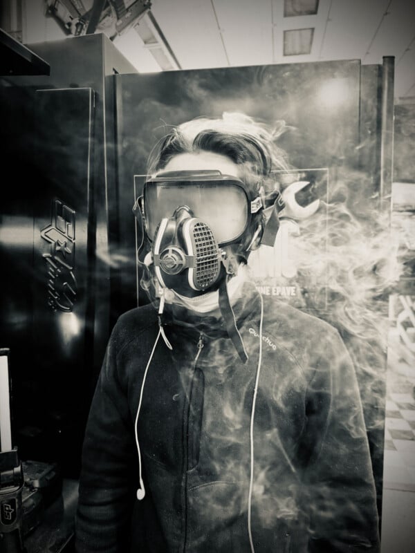 A person wearing a protective mask and goggles stands amid swirling smoke, dressed in a dark jacket with earphones dangling. The black-and-white image has an industrial, gritty atmosphere.