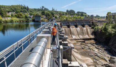 A person in orange safety clothes stands on a dam. On one side is a lake. On the other are dry spillways that lead to a rocky riverbed.