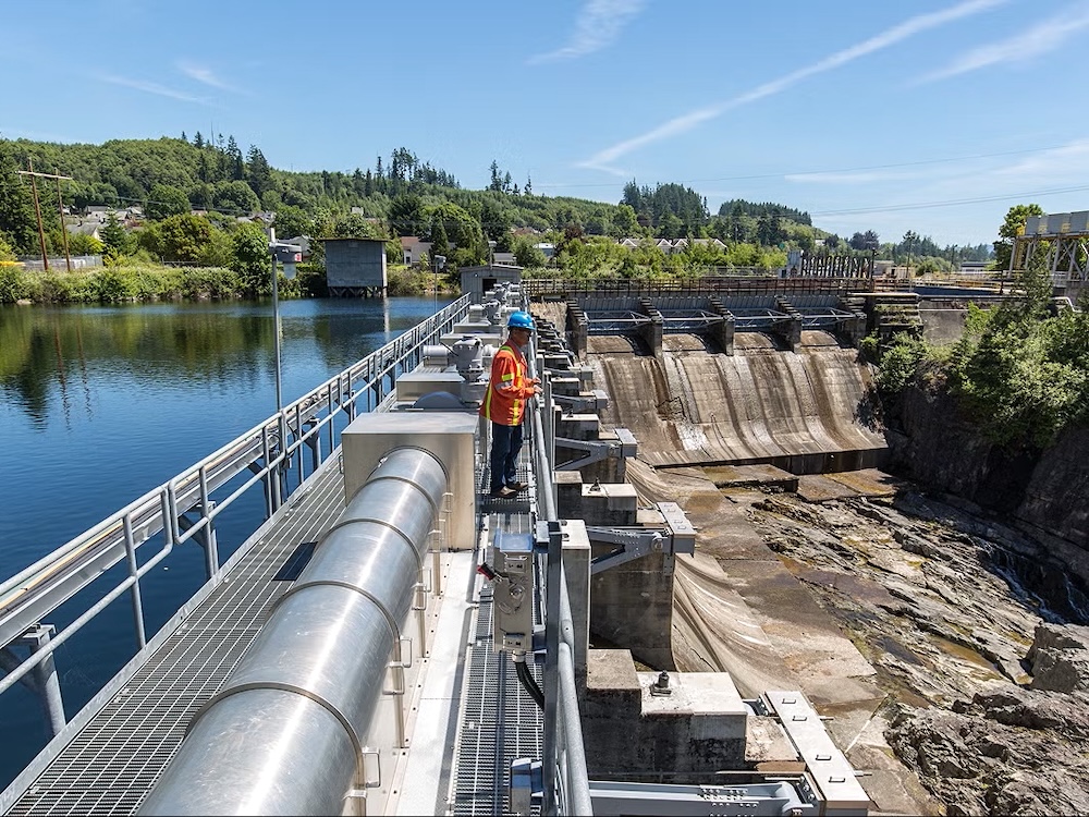 A person in orange safety clothes stands on a dam. On one side is a lake. On the other are dry spillways that lead to a rocky riverbed.