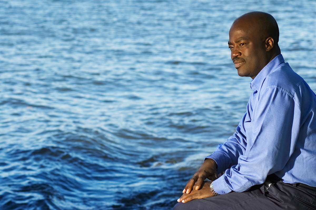 A bald man with dark skin tone wears a blue collared shirt and sits in front of the ocean.