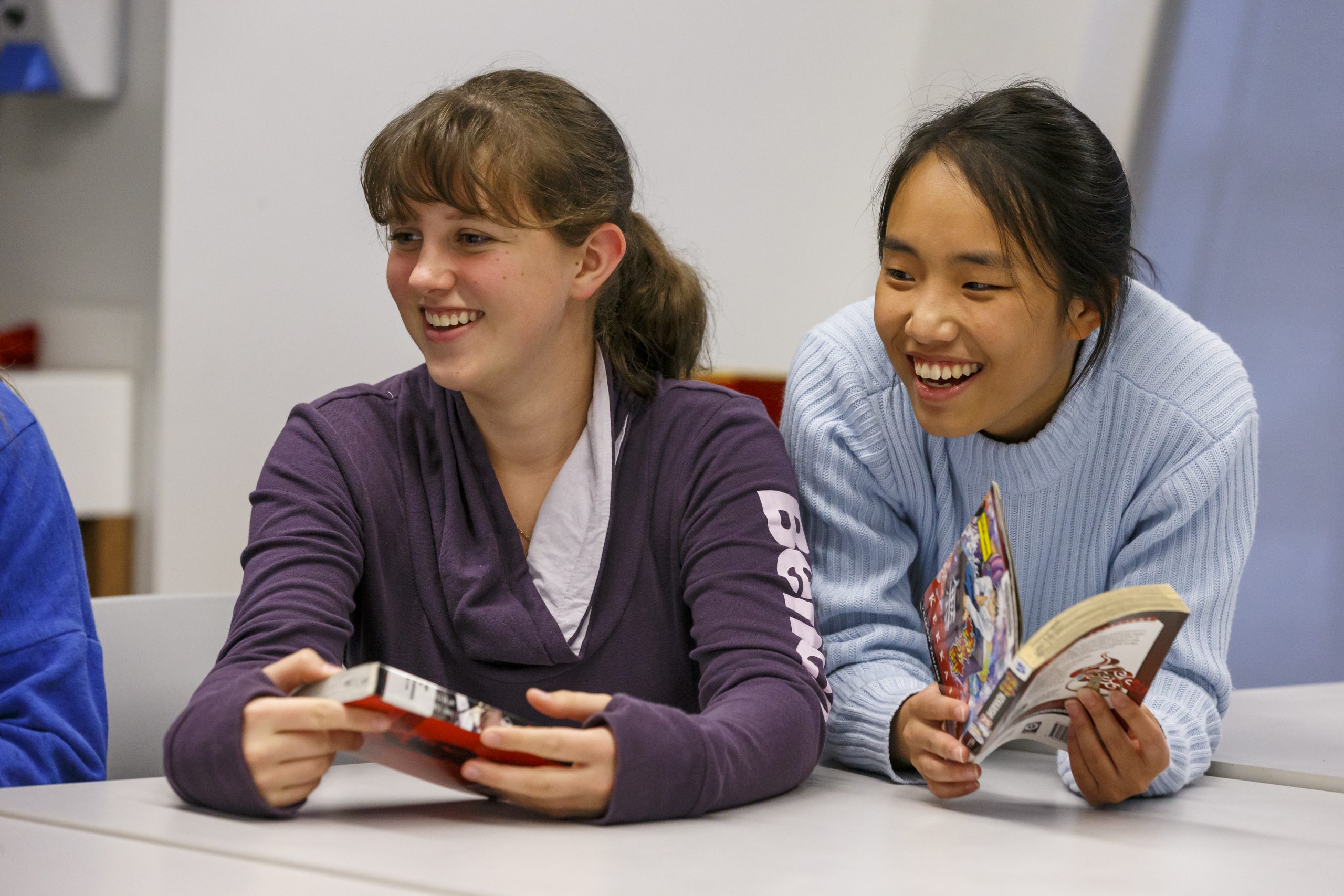 Two library visitors share and discuss graphic novels at a library table, highlighting programs that encourage reading, learning, and community engagement for teens.