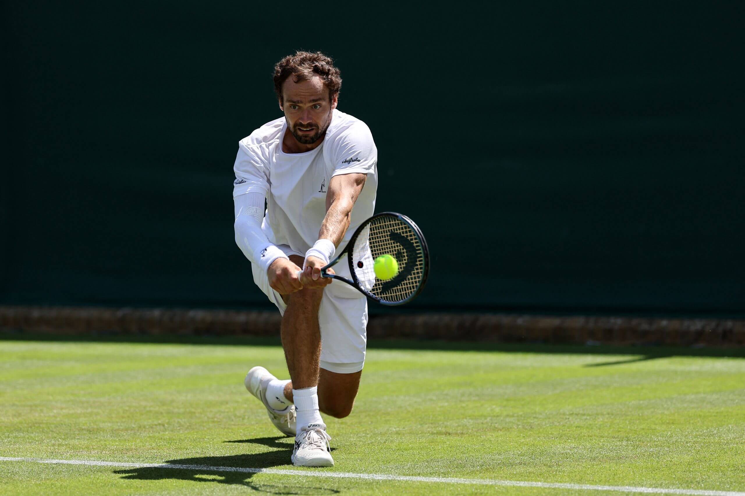 Roman Safiullin hits a two-handed backhand low on a grass tennis court.