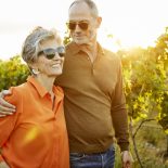 Happy woman walking with male partner in vineyard during sunset. Elderly couple are touring in field against sky. They are enjoying during weekend.