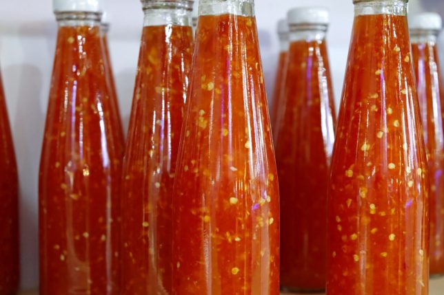 Rows of Glass Bottles Filled with Spicy and Sweet Thai Chili Sauce, Showcasing the Vibrant Red Color and Visible Flecks of Chili Seeds and Garlic, Ready for Sale in a Market or Grocery Store