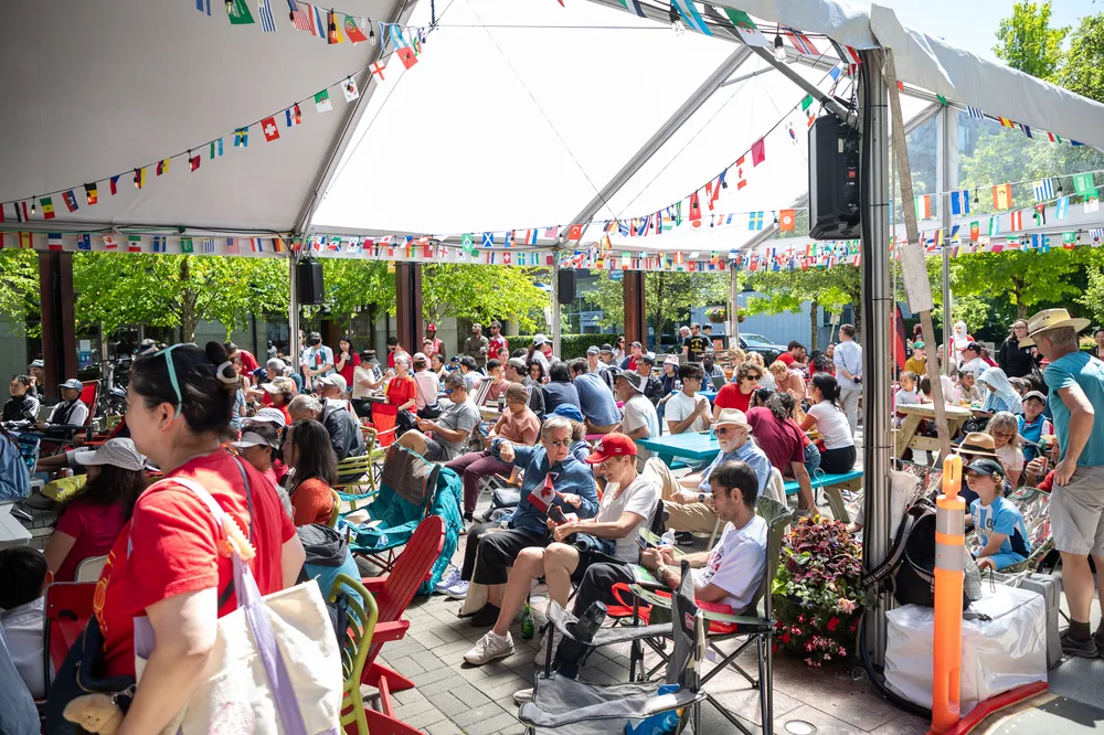A large group of people gather outside, under a tent, to watch a world cup match with friends and family.