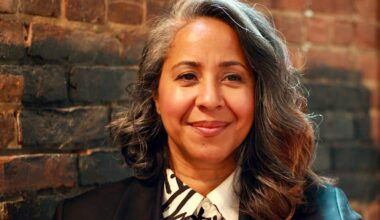 A middle-aged woman with long curly grey and brown hair smiles warmly against a background of a brick wall.