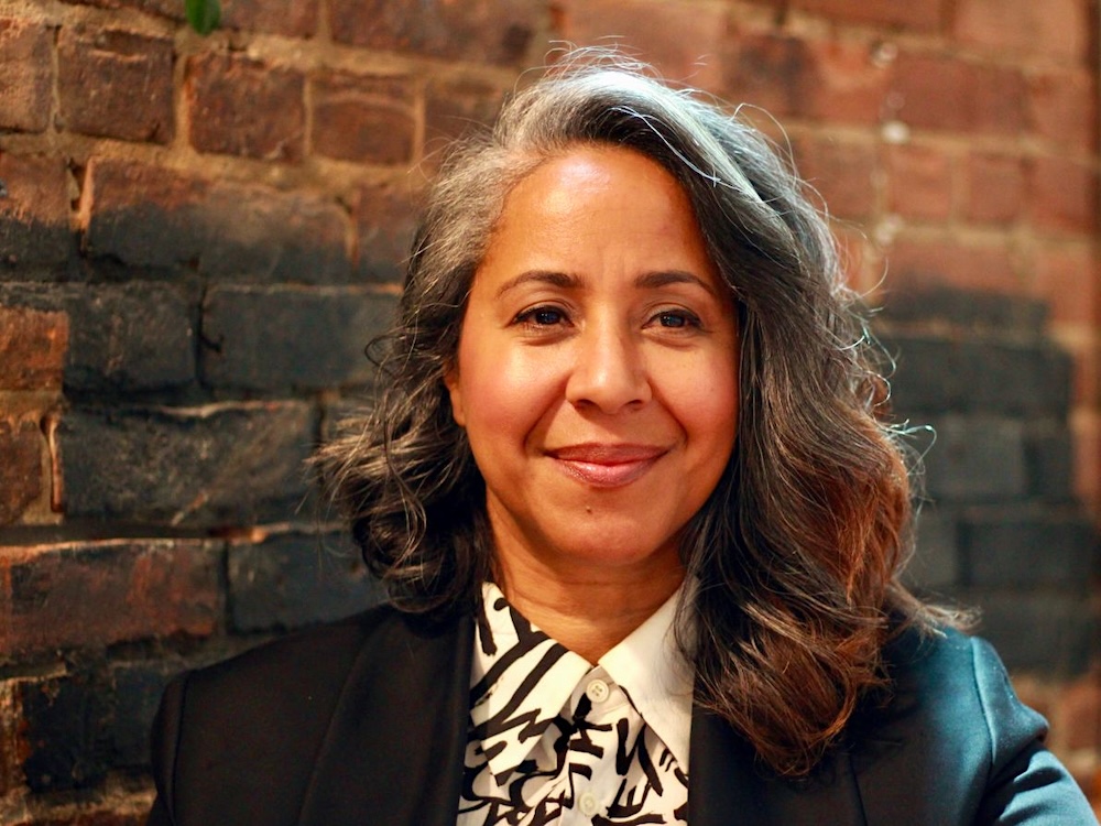 A middle-aged woman with long curly grey and brown hair smiles warmly against a background of a brick wall.