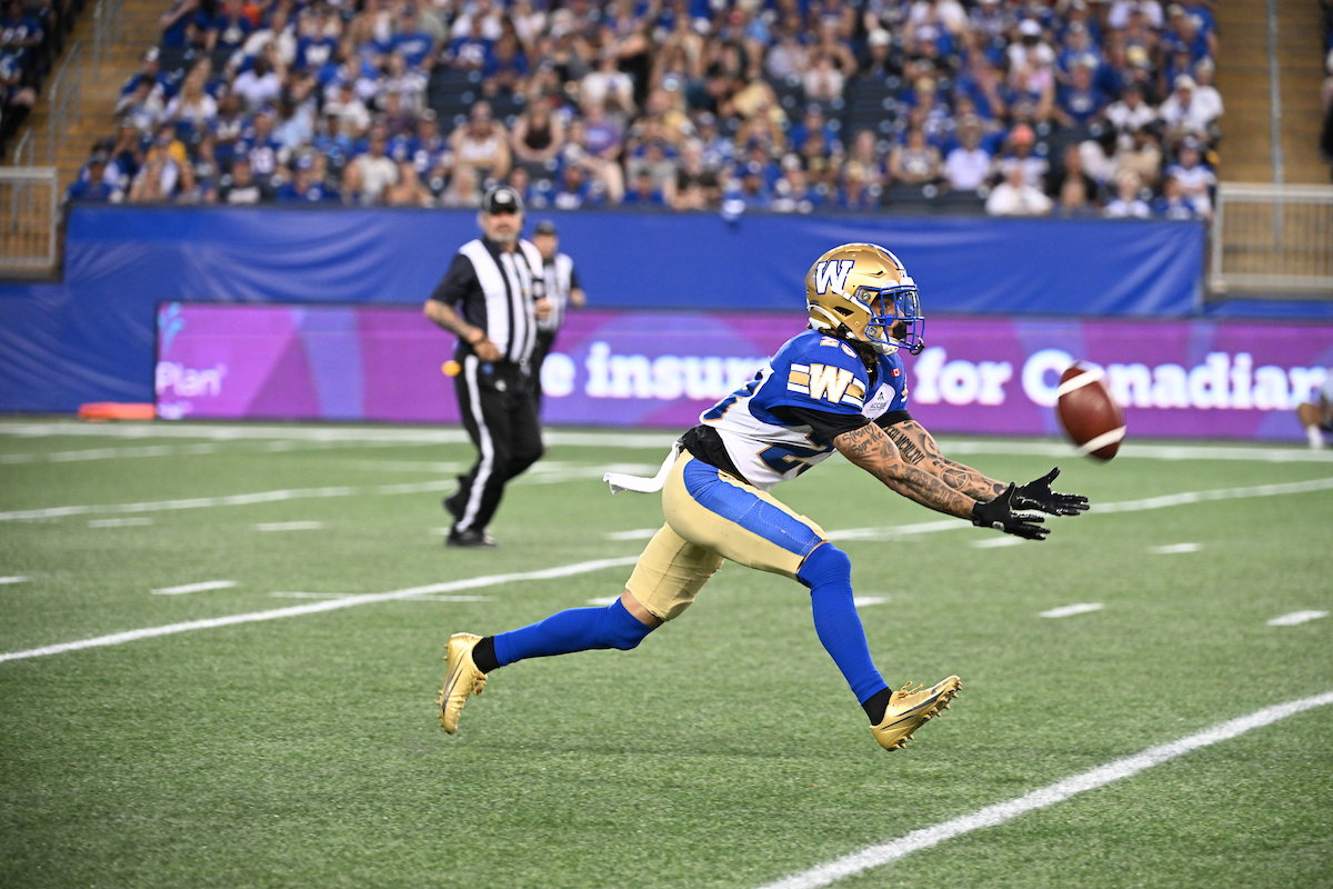 A football player in a blue and gold uniform leaps to catch a pass during a game, with a referee and spectators in the background.