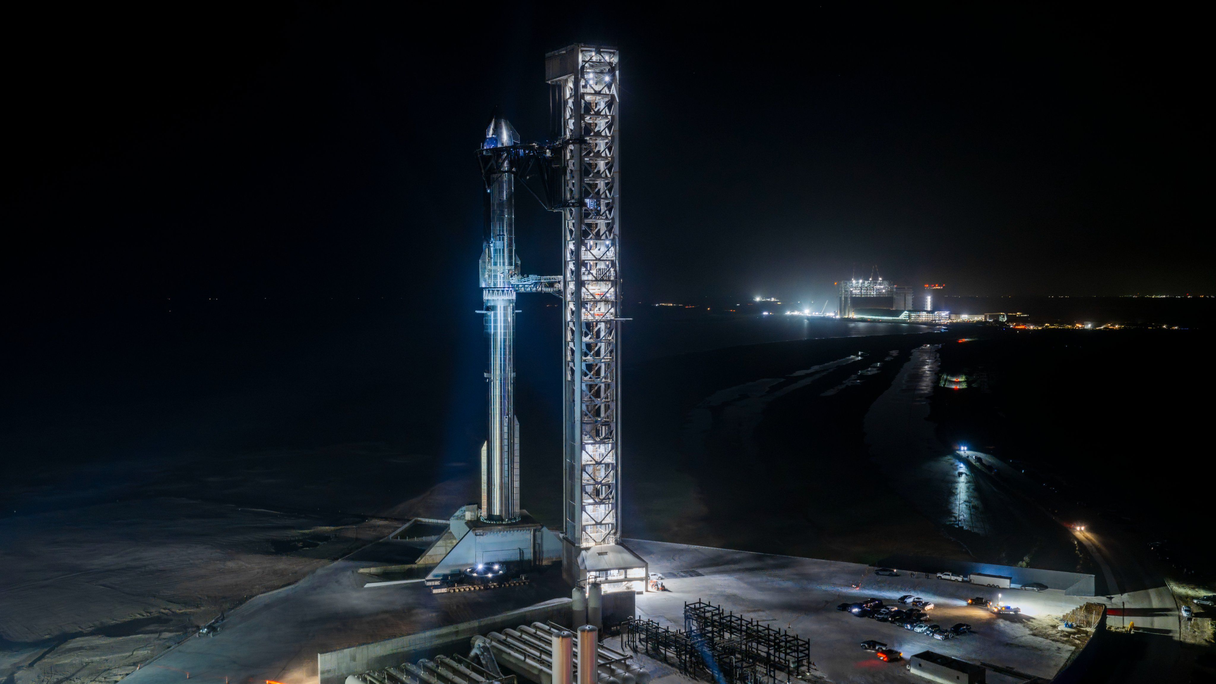 A giant steel rocket stands next to a launch tower lit by spotlights in the dark of night.