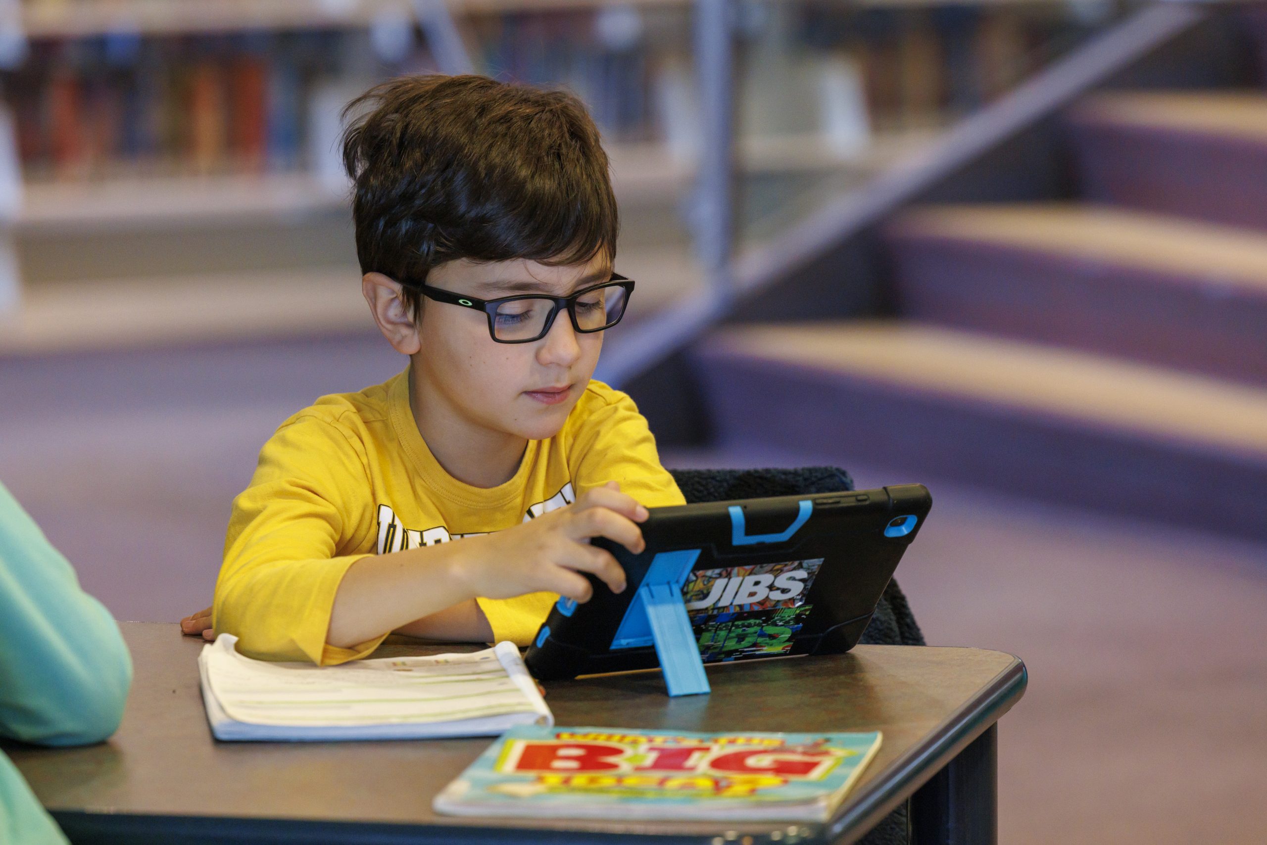 A child library visitor uses a tablet alongside print materials at a library table, demonstrating access to both digital and physical collections.