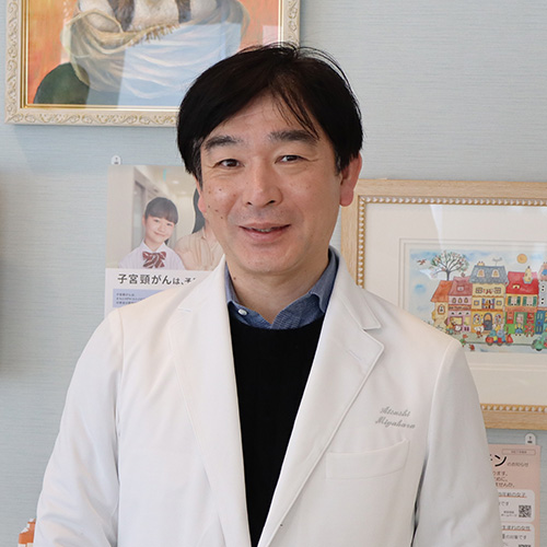 An Asian man wearing a white lab coat with a black blazer and black hair looks at the camera