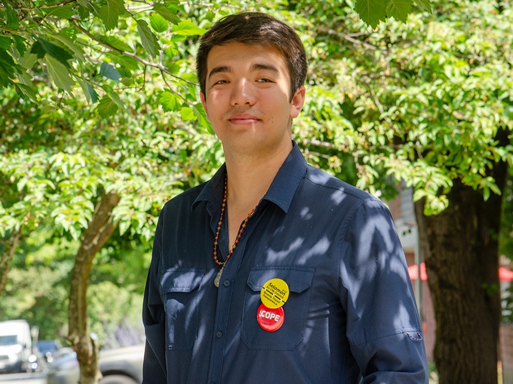 A young person with short black hair stands in an outdoor setting.