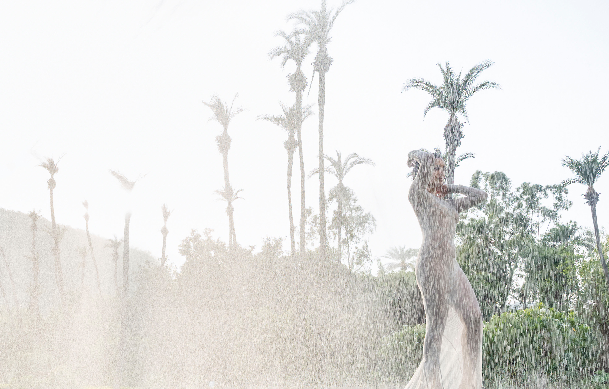 A woman standing in a transparent dress in front of palms in the ran