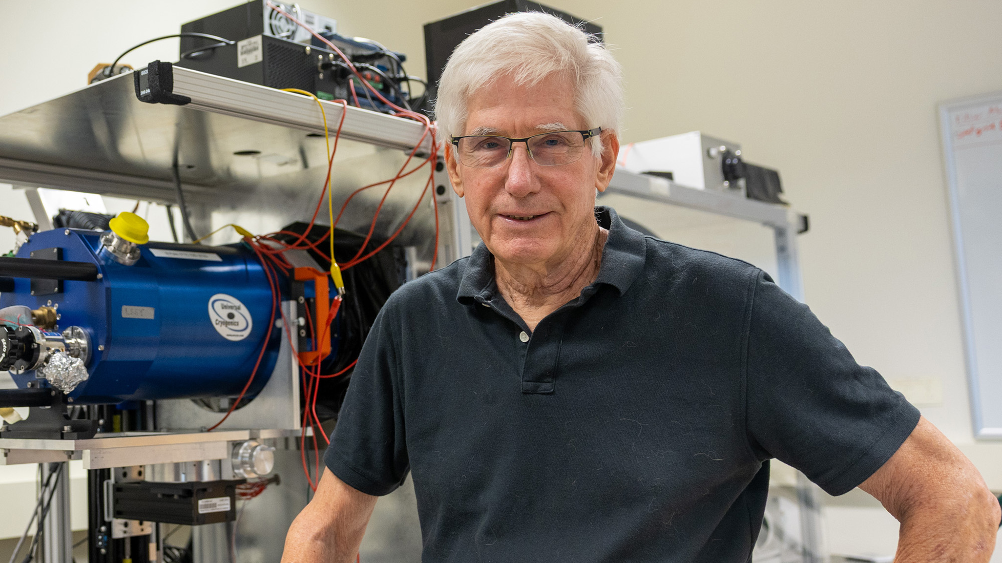 A man with white hair, glasses and a black polo shirt stands in front of some laboratory equipment