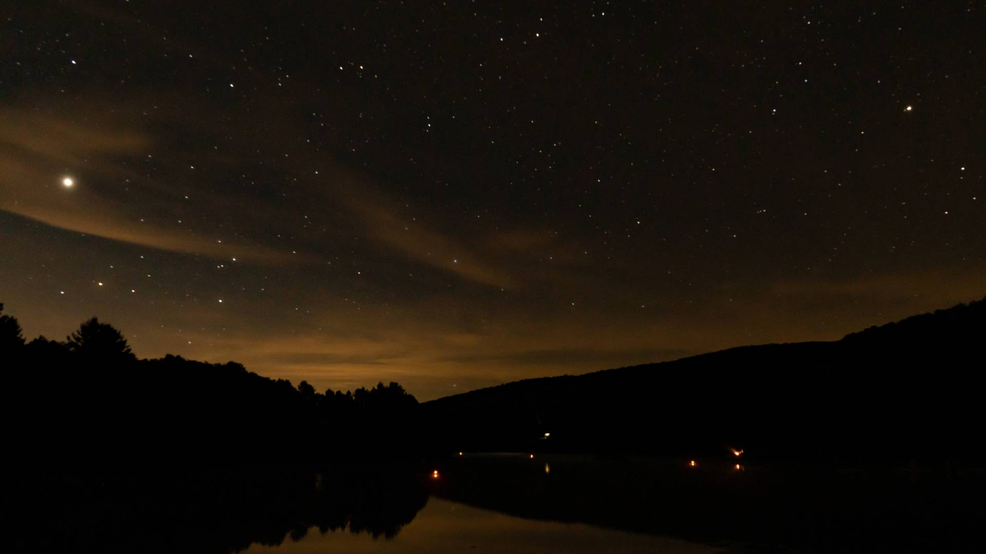 A star-filled night sky reflected over a still lake, with distant hills silhouetted against a faint orange glow and wispy clouds on the horizon.