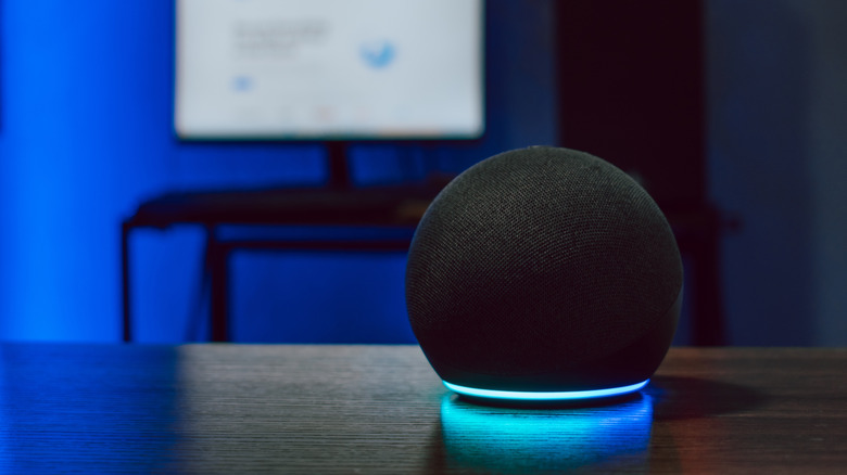 An Amazon Echo Dot placed on a wooden surface in a room bathed in blue light, with a blurry monitor in the background