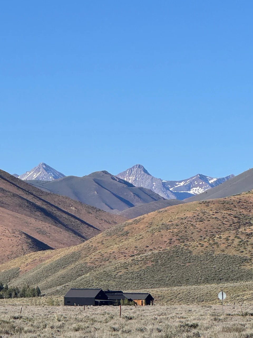 A mountain landscape with sagebrush hills, snow-capped peaks, and a ranch building under a clear blue sky.