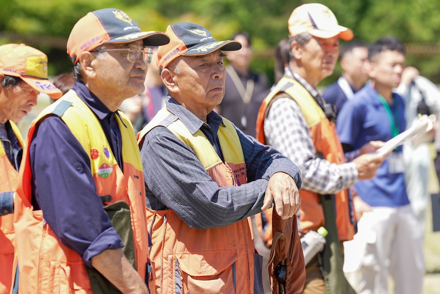 men in caps and orange vests stand together listening to a talk