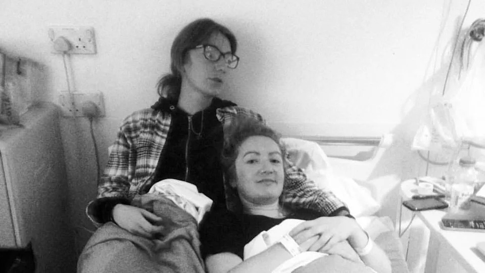 A black and white photo of Lowri in hospital in September 2015 waiting for scans with her sister. Lowri is lying in bed and her sister had her arm around her. Both have their hair tied back. Lowri's sister is wearing a hoodie.