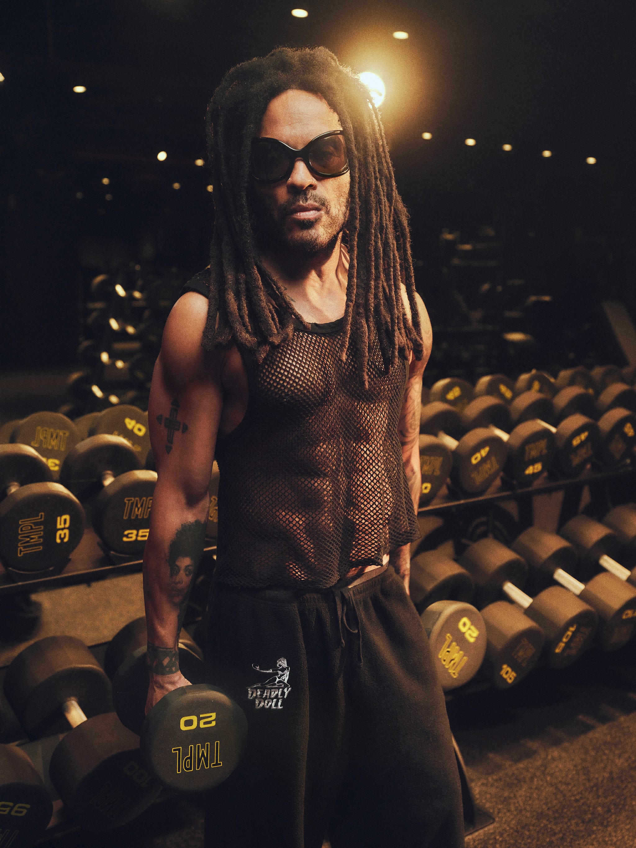 Person with dreadlocks and sunglasses holding a dumbbell in a gym.