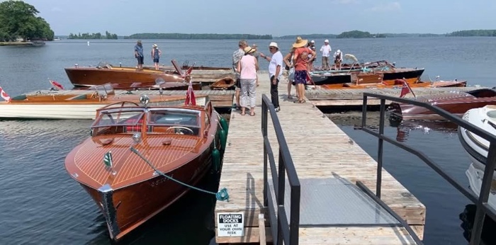 people in summer clothes stand on a wooden dock beside several polished wooden boats moored to the pier on a calm lake.