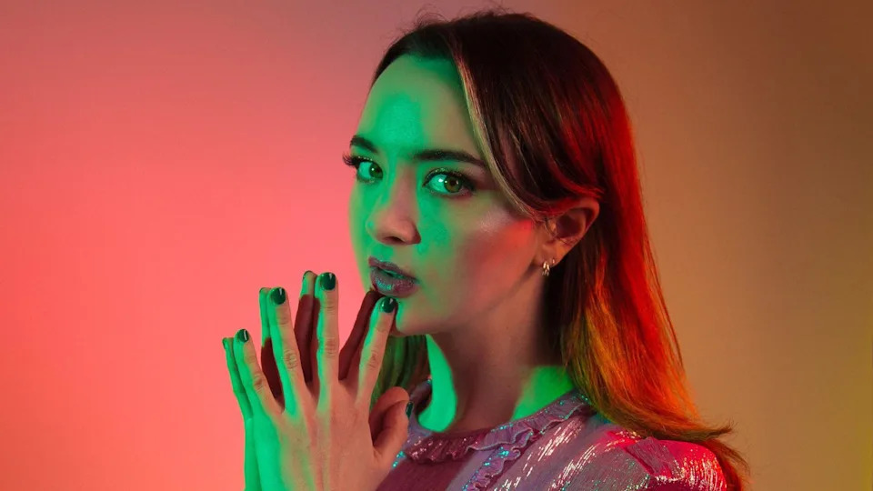 Fern Brady posing for a promotional picture, lit in red and green artistic light. She has long dark hair and green eyes. She is wearing a pink sparkly dress and is holding her hands in a prayer gesture under her chin. She is looking sideways at the camera.